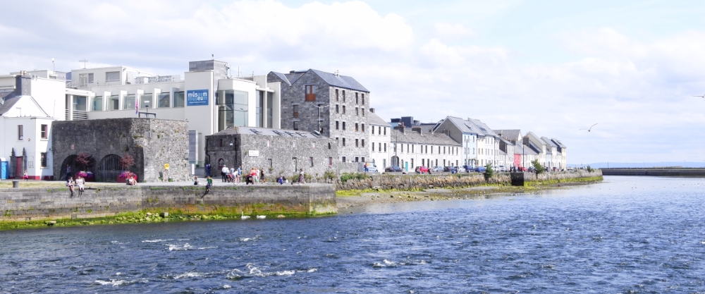 View of Galway's seafront promenade with the grey stone Spanish Arch and a row of traditional houses facing the River Corrib.